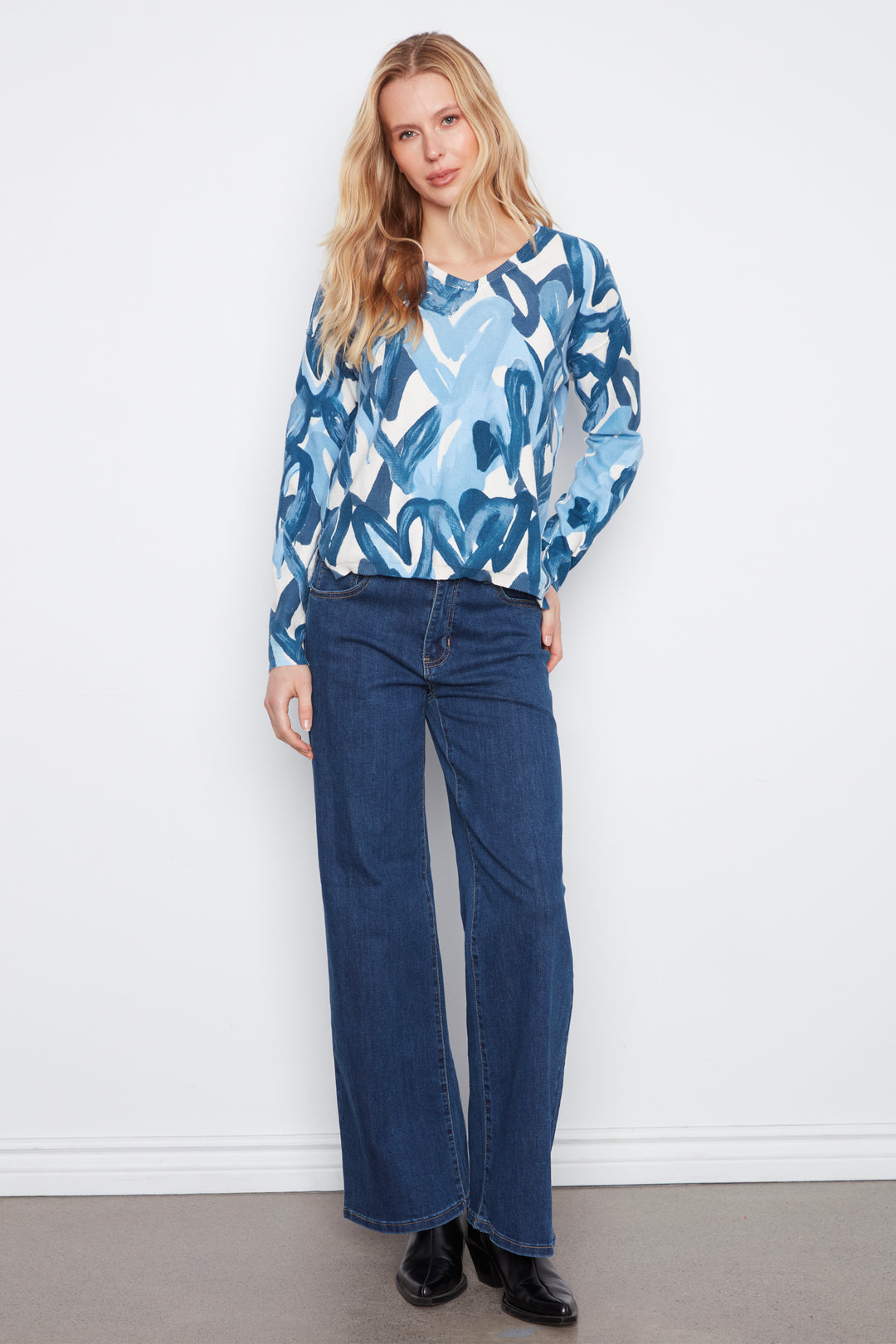 A woman stands against a white wall wearing a light blue and white abstract print long-sleeve blouse with dark blue wide-leg jeans and black shoes.