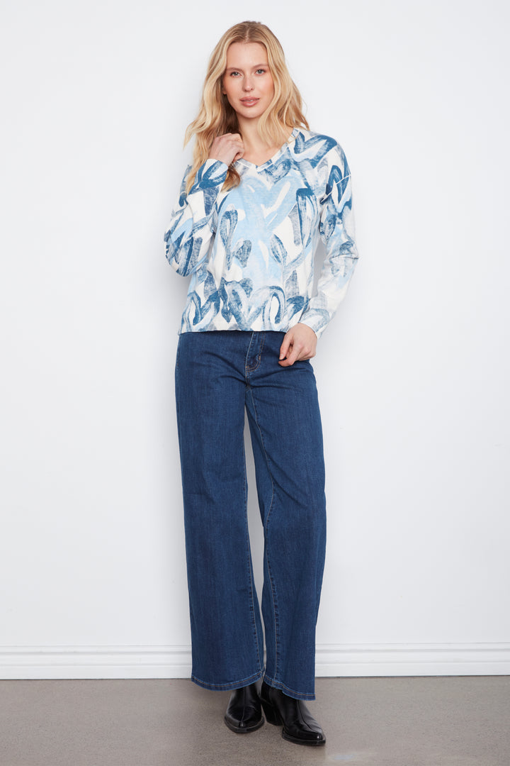 A woman stands against a white wall wearing a light blue and white abstract print long-sleeve blouse with dark blue wide-leg jeans and black shoes.