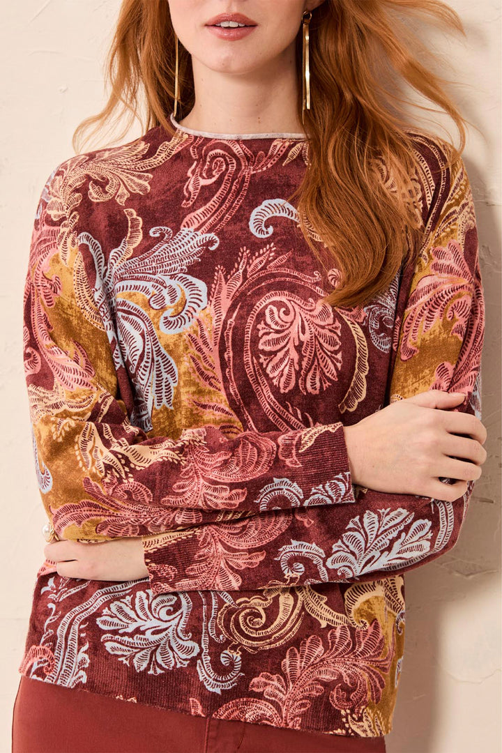 A woman wearing a long-sleeve crewneck top with an intricate paisley and floral pattern in rust red, mustard yellow, dusty pink, and white. She is leaning against a light beige textured wall.