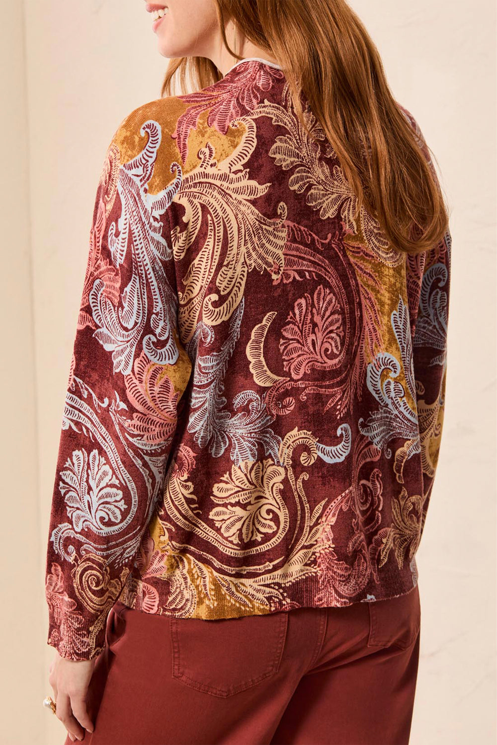 A woman wearing a long-sleeve velvet top with an intricate multicolor paisley pattern in burgundy, gold, and silver, paired with rust-colored pants.