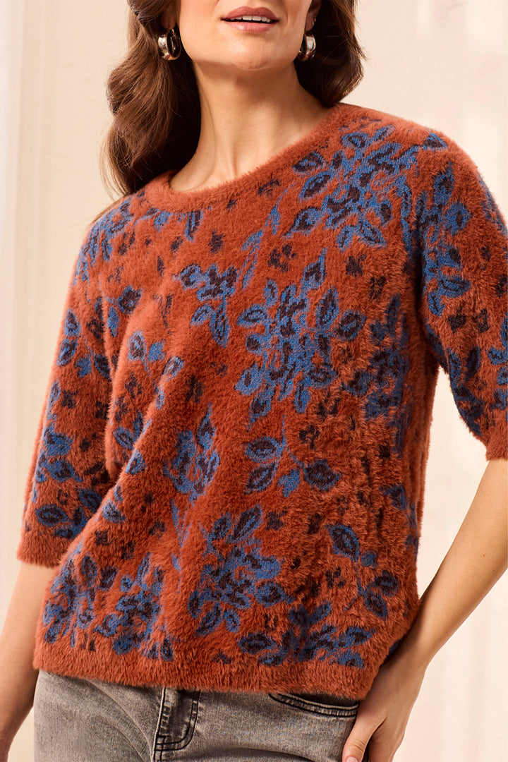 A woman wearing a fuzzy short-sleeve knit sweater with a rust-orange base and blue floral pattern, paired with gray jeans.