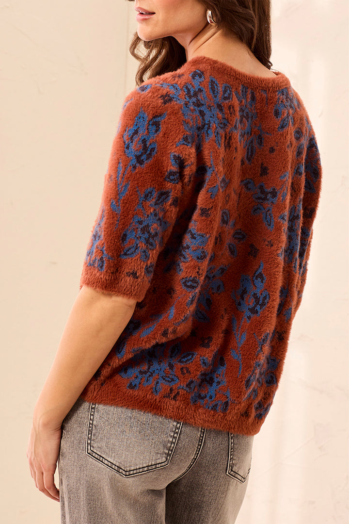 A woman wearing a fuzzy short-sleeve knit top in rust orange with a blue floral pattern, paired with gray jeans, shown from a side-back angle against a plain beige background.