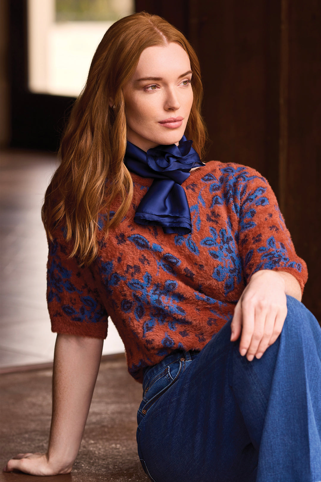A woman with long red hair sits on a stone floor wearing a rust-orange and blue patterned knit sweater, a dark blue satin neck scarf, and blue jeans in a dimly lit interior setting.