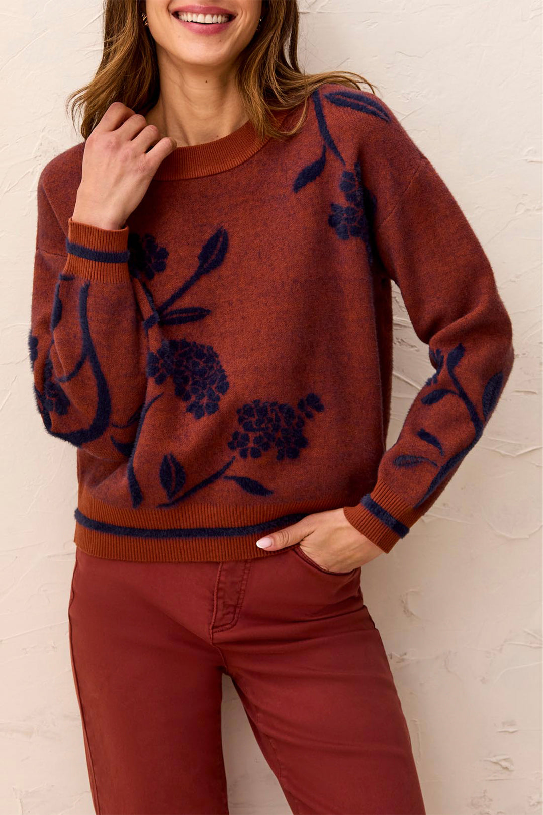 A woman wearing a rust-brown crewneck sweater with a navy blue floral pattern, paired with rust-brown pants, standing against a textured off-white wall.