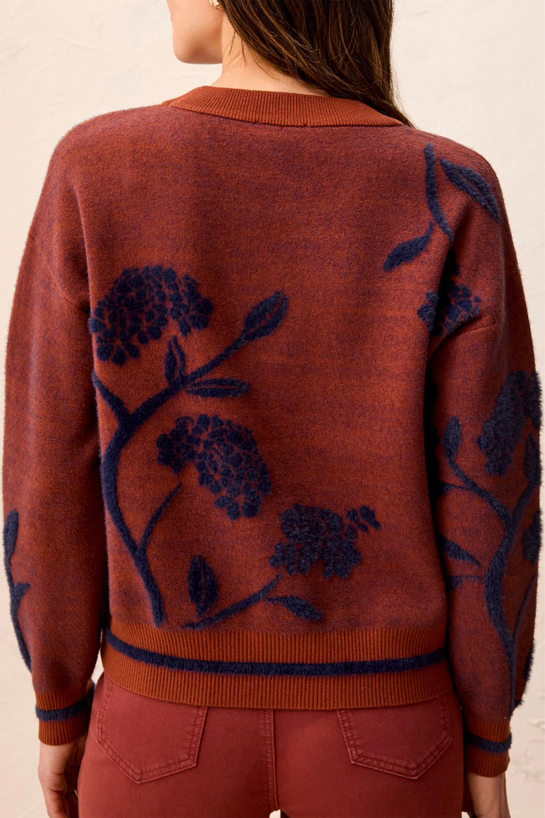 A woman viewed from the back wearing a rust-orange knit sweater with dark navy floral embroidery and matching rust-orange pants against a light beige background.