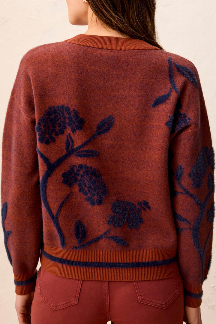 A woman viewed from the back wearing a rust-orange knit sweater with dark navy floral embroidery and matching rust-orange pants against a light beige background.
