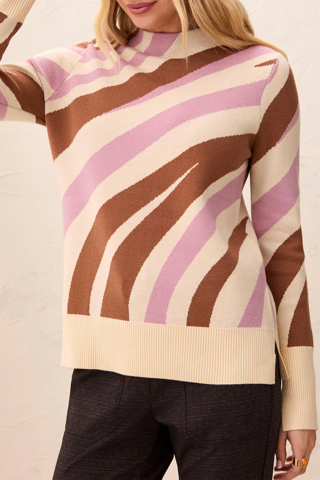 A woman wearing a knit sweater with wavy diagonal stripes in brown, light pink, and cream, paired with dark charcoal pants.