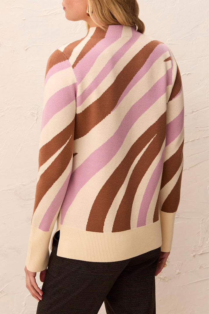 A woman shown from the back wearing a knit sweater with bold diagonal stripes in brown, light pink, and cream, paired with dark charcoal pants against a textured off-white wall.