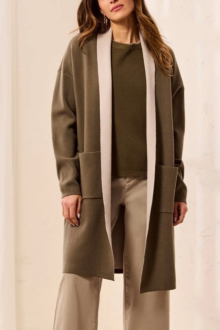 A woman wearing a long olive green open-front cardigan with contrasting light beige trim, paired with a dark olive sweater, beige pants, and gold hoop earrings against a neutral cream background.