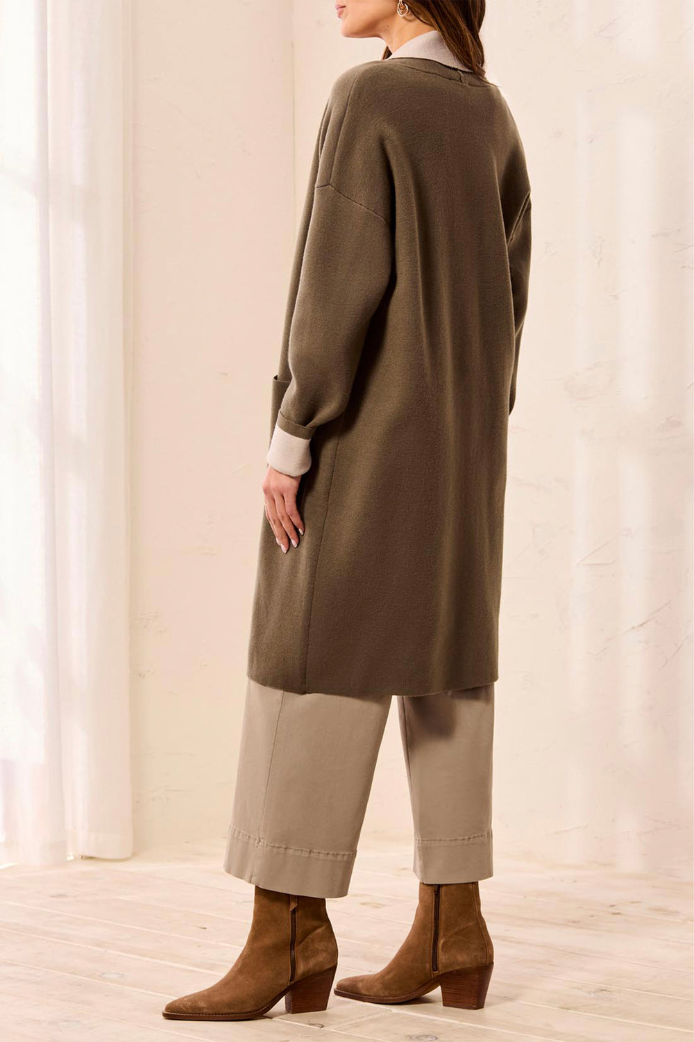 A woman shown from the back wearing a long olive green knit cardigan over a cream shirt, paired with beige cropped pants and brown suede ankle boots in a bright indoor setting.