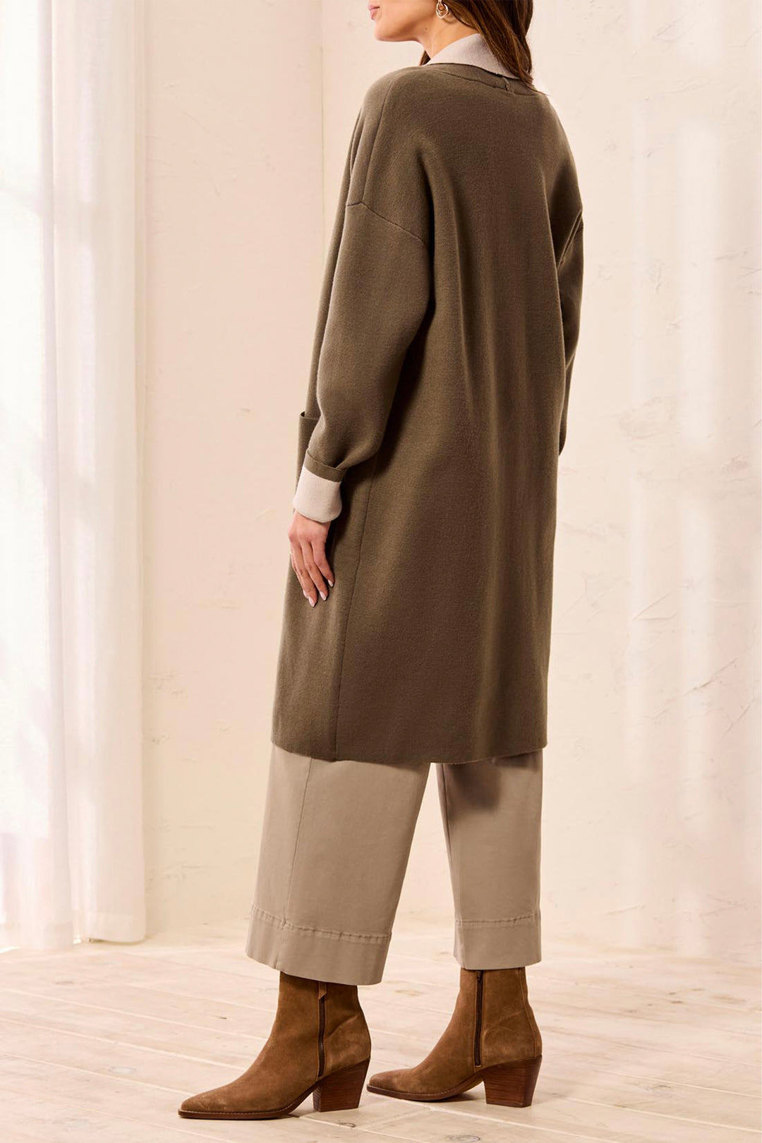 A woman shown from the back wearing a long olive green knit cardigan over a cream shirt, paired with beige cropped pants and brown suede ankle boots in a bright indoor setting.