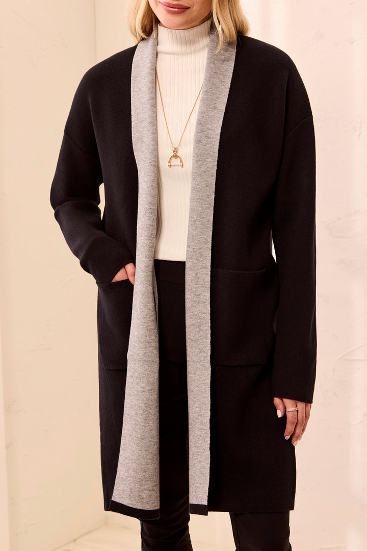 A woman wearing a long black open-front cardigan with contrasting light grey trim against a neutral cream background.