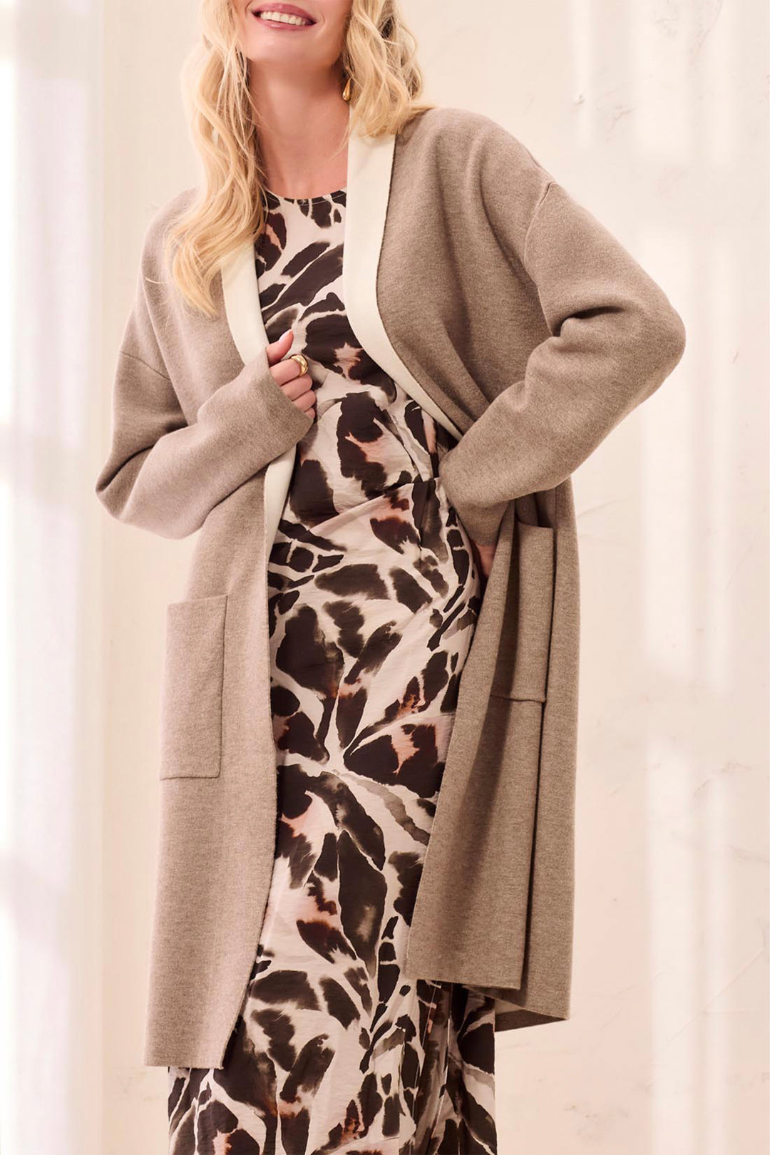 A woman wearing a taupe open-front cardigan over a brown, black, and beige animal-print dress, standing against a soft white curtain background.