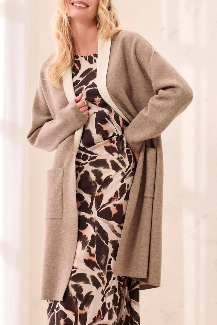A woman wearing a taupe open-front cardigan over a brown, black, and beige animal-print dress, standing against a soft white curtain background.