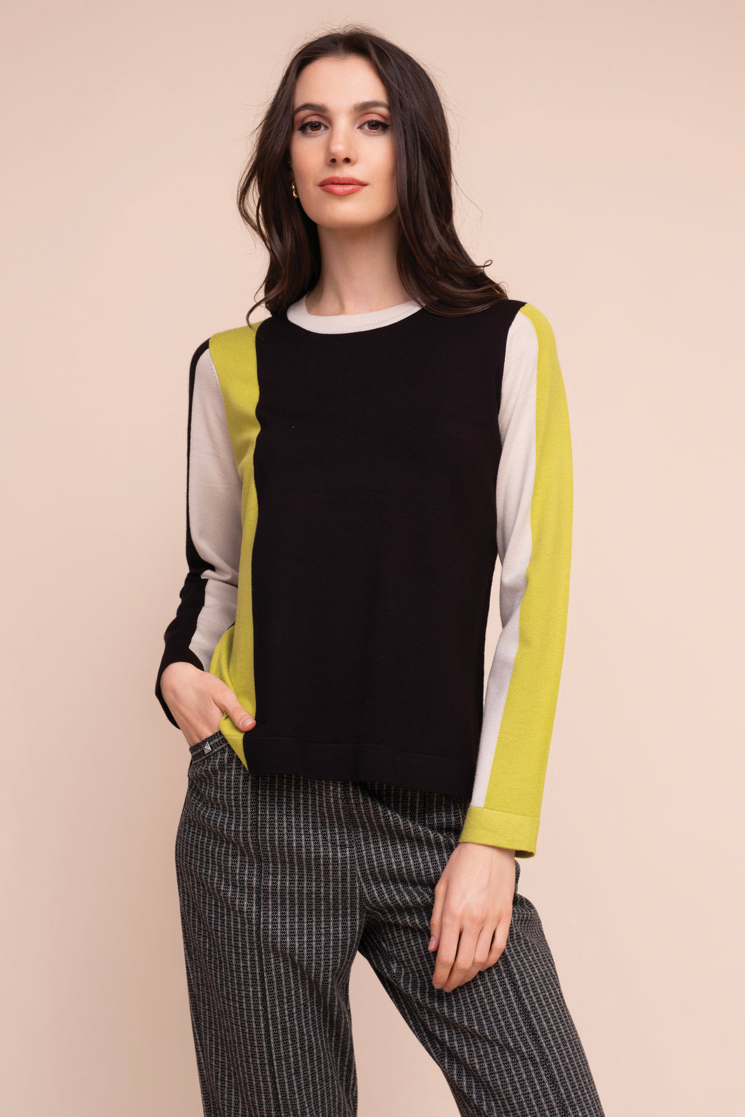 A woman modeling a color-block knit sweater with a black body, lime green side panels, and white sleeves, paired with gray pinstriped trousers, against a solid beige background.