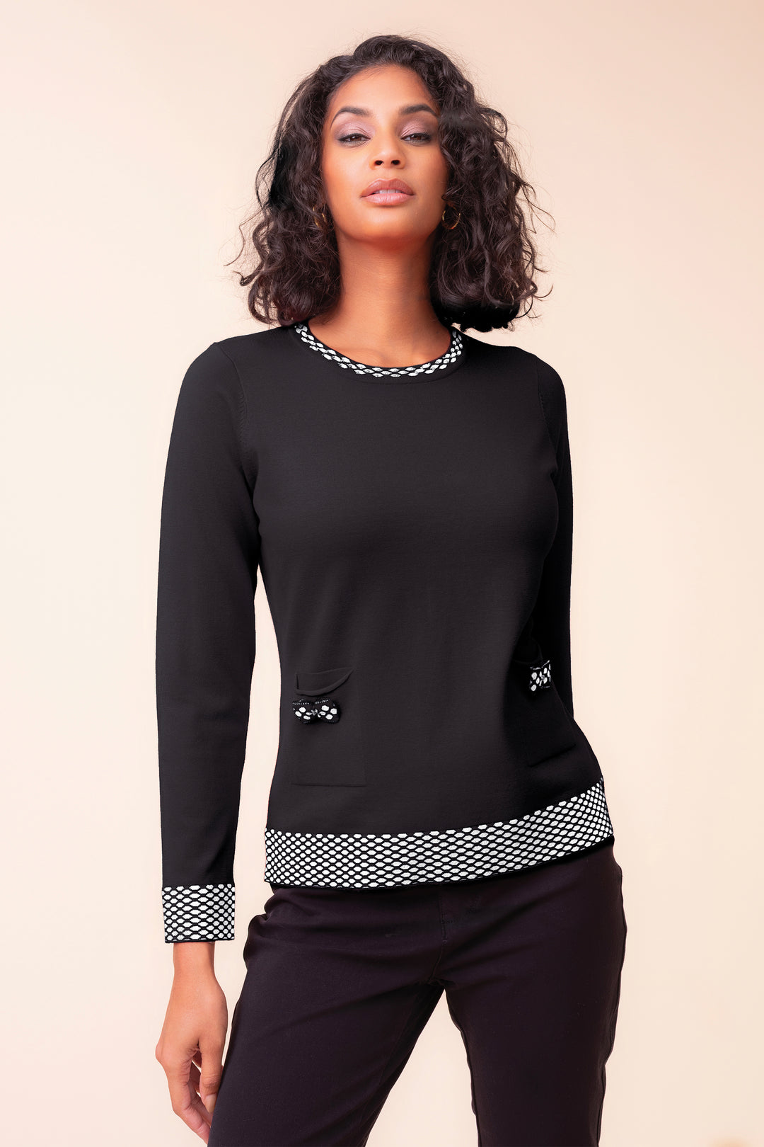 A woman modelling a black sweater with geometric pattern on the collar, hem and cuffs, standing in front of a beige background. 