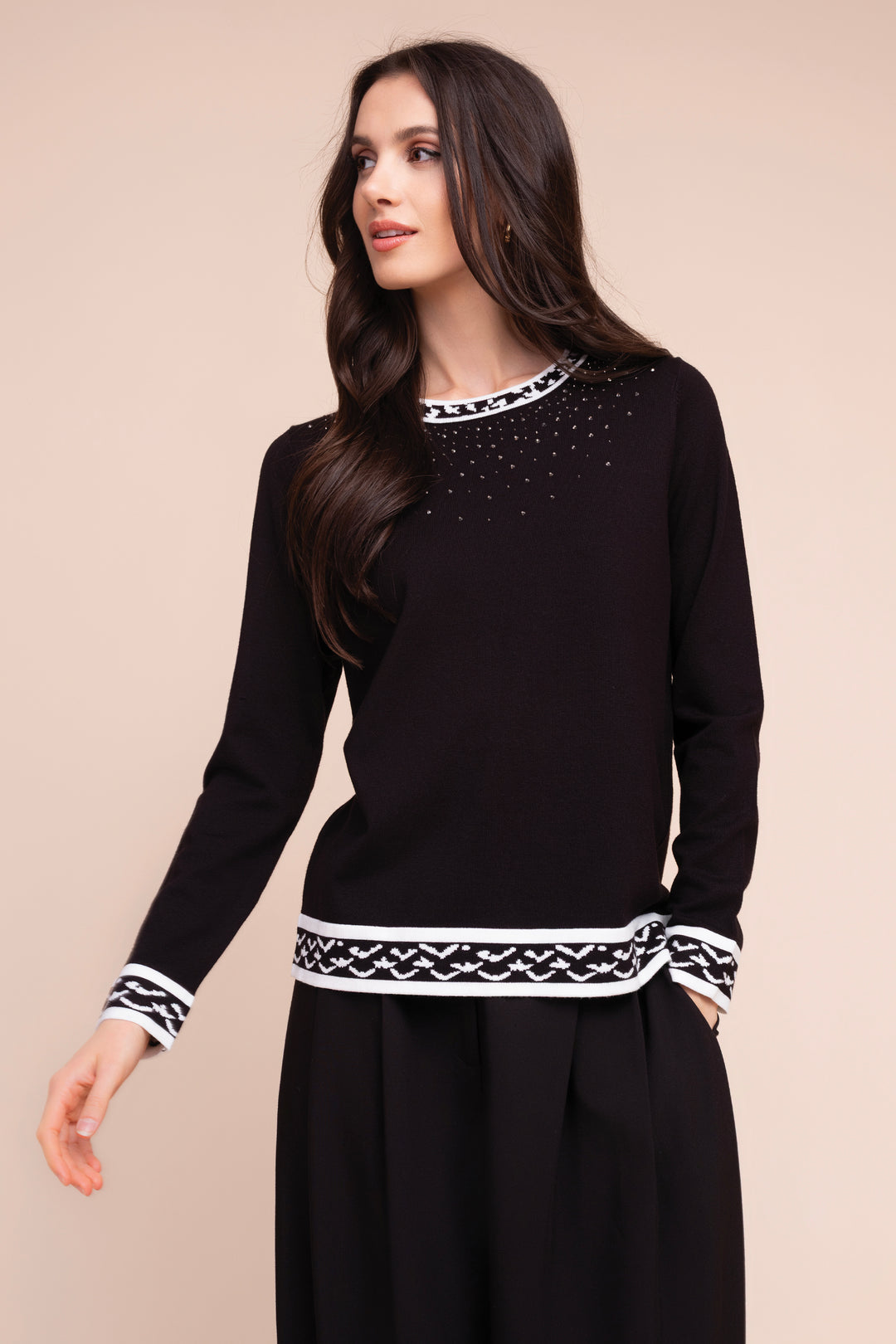 A woman modeling a black long-sleeve knit top with white geometric trim at the crew neck, cuffs, and hem, featuring small silver rhinestone embellishments on the chest, set against a solid beige background.