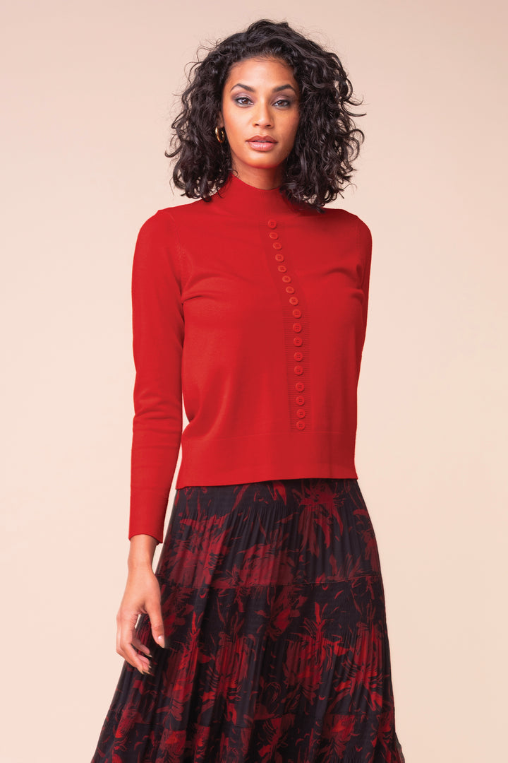 A woman modeling a red mock-neck long-sleeve knit top with a button-down front, paired with a dark red and black patterned pleated skirt, against a solid beige background.