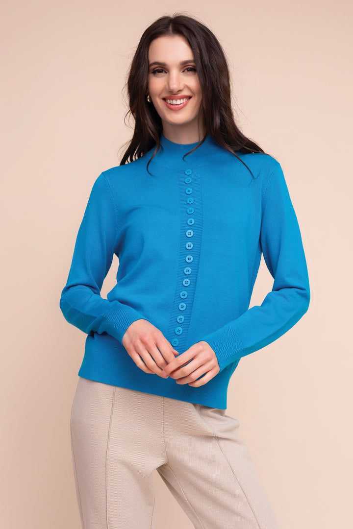 A woman modeling a bright blue mock-neck knit cardigan sweater with a button-down front, paired with beige trousers, against a solid beige background.