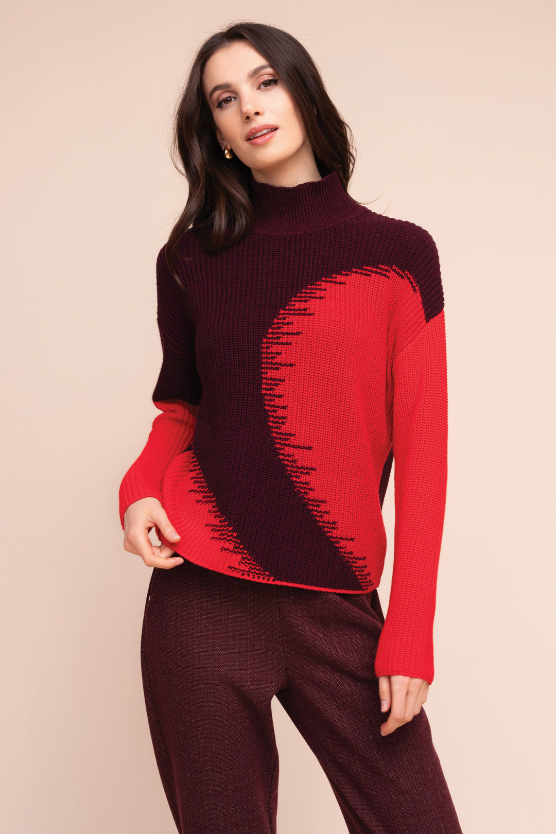 A woman modeling a long-sleeve mock neck knit sweater with a color-blocked design in deep burgundy and bright red, paired with matching burgundy pants, against a solid beige background.