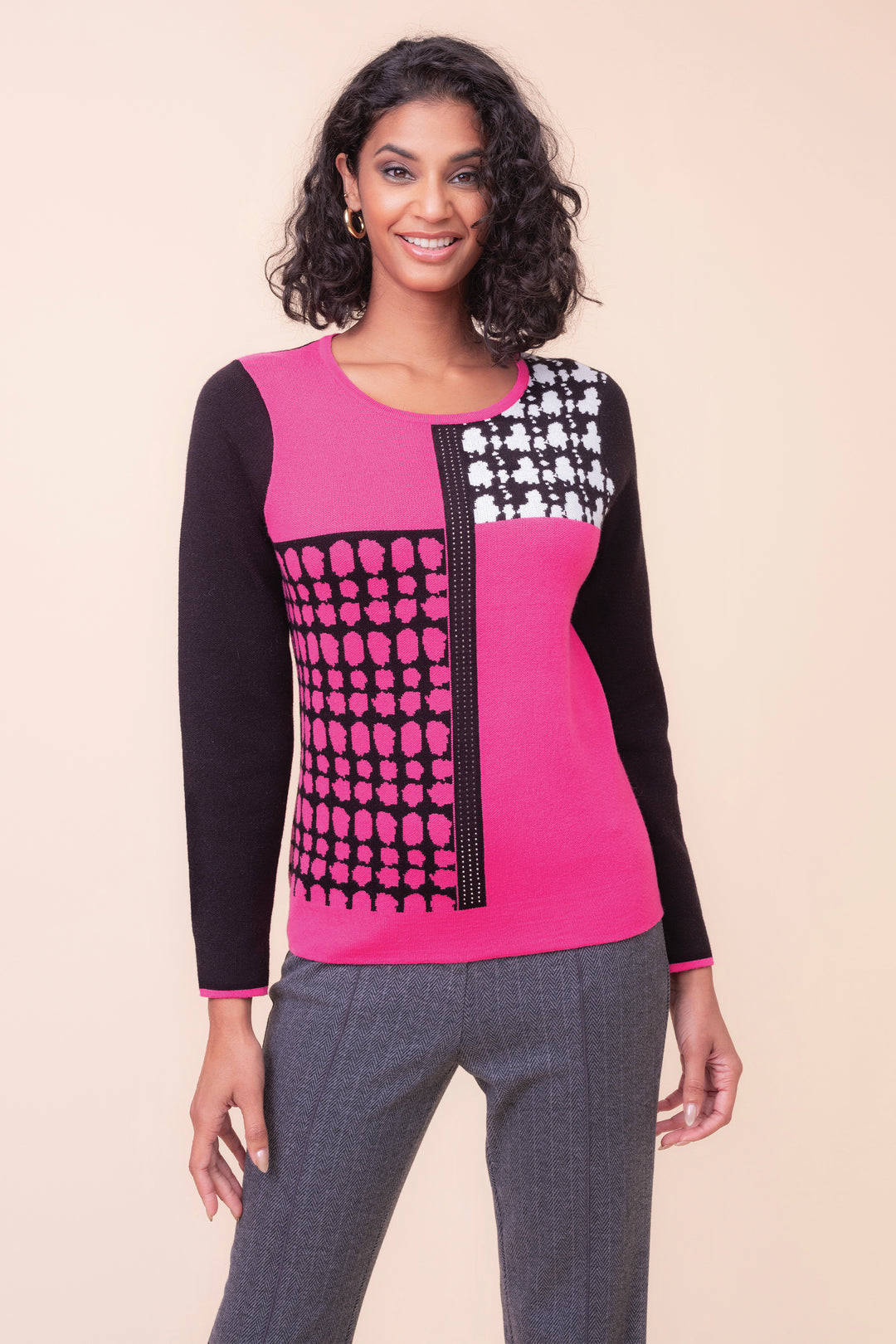 A woman modeling a bright pink and black long-sleeve knit top with a geometric patterned chest panel, paired with gray pants, against a solid beige background.