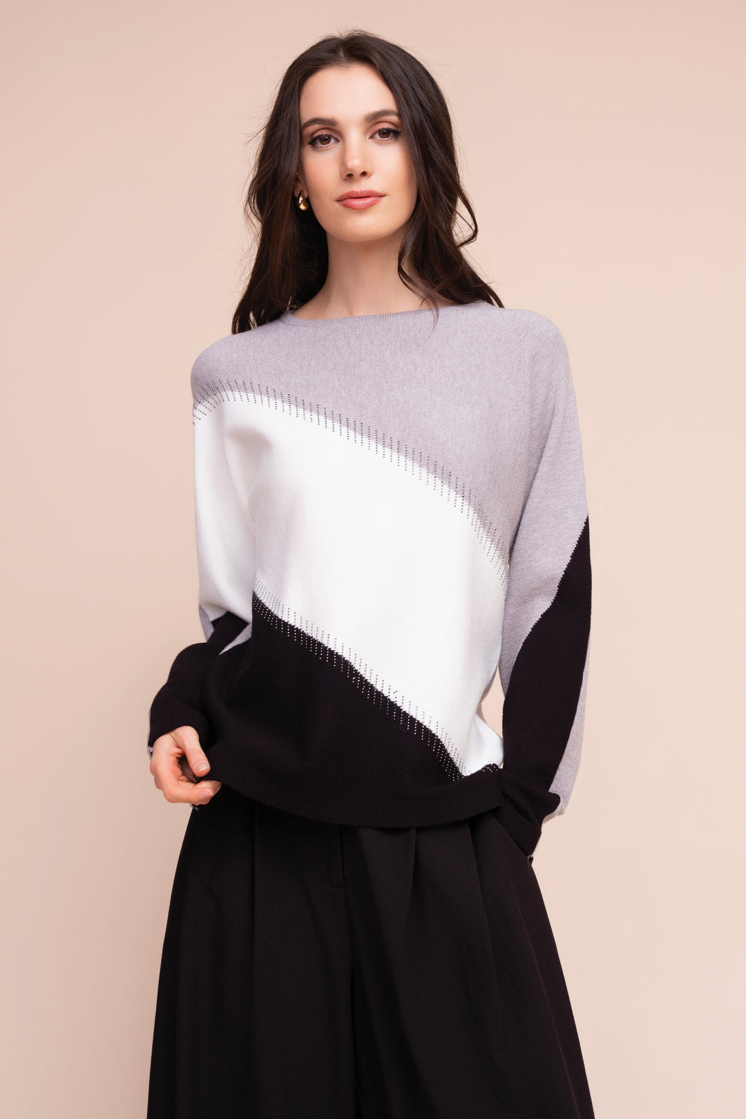 A woman modeling a color-block knit sweater with a wide boat neckline, featuring light gray, white, and black panels, paired with a black skirt against a solid beige background.