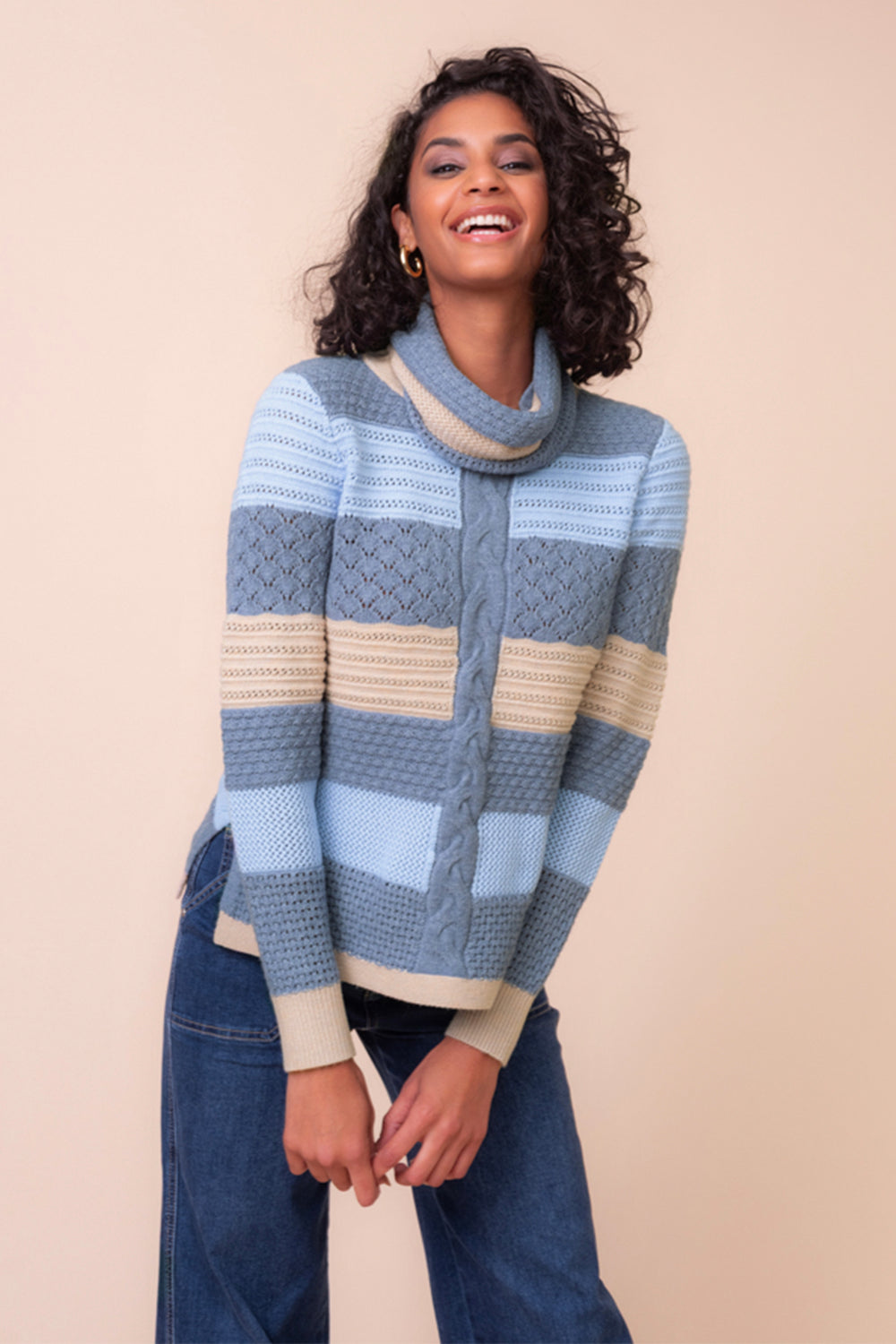 A woman modeling a chunky knit cowl-neck sweater with horizontal stripes in blue, taupe, and denim blue, paired with jeans, against a solid pale peach background.