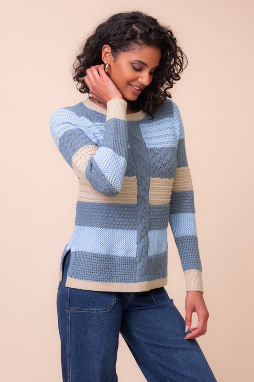 A woman modeling a chunky knit sweater with horizontal stripes in blue, taupe, and denim blue, paired with jeans, against a solid pale peach background.