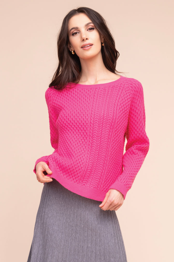 A woman modeling a bright pink long-sleeve knit sweater with a textured cable pattern, paired with a gray pleated skirt, against a solid beige background.