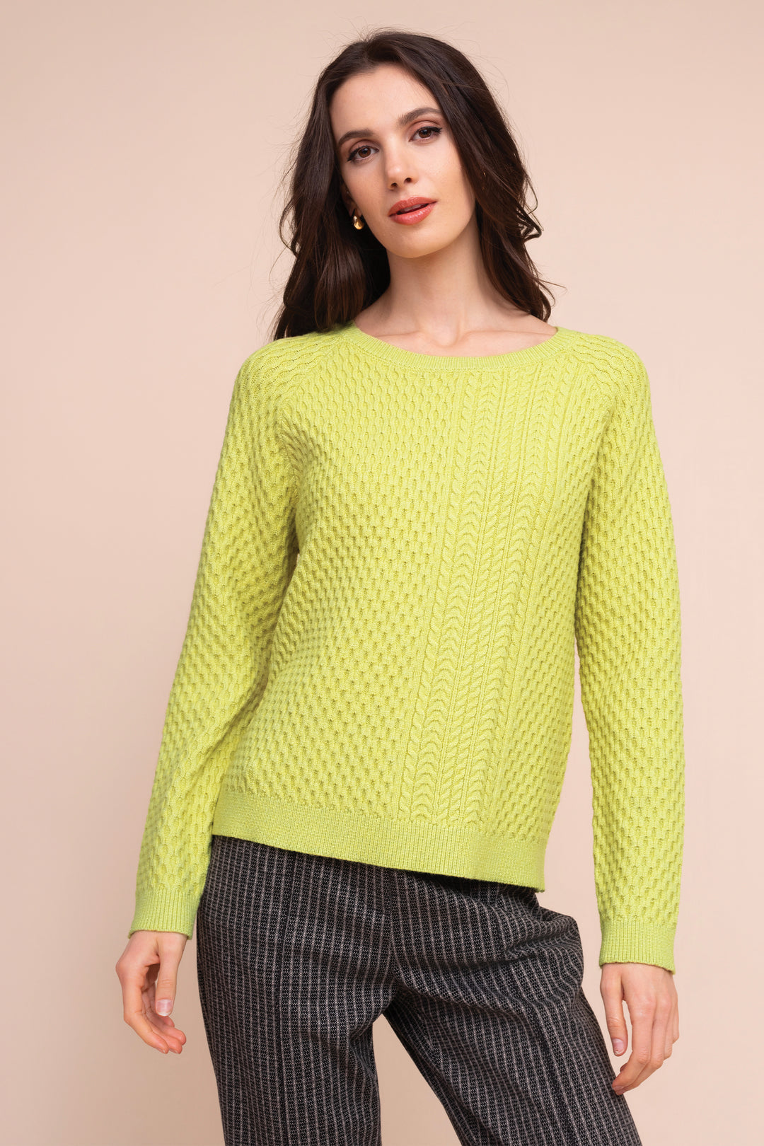 A woman modeling a bright lime green knit sweater with a textured cable pattern, paired with dark gray pinstriped trousers, against a solid pale peach background.