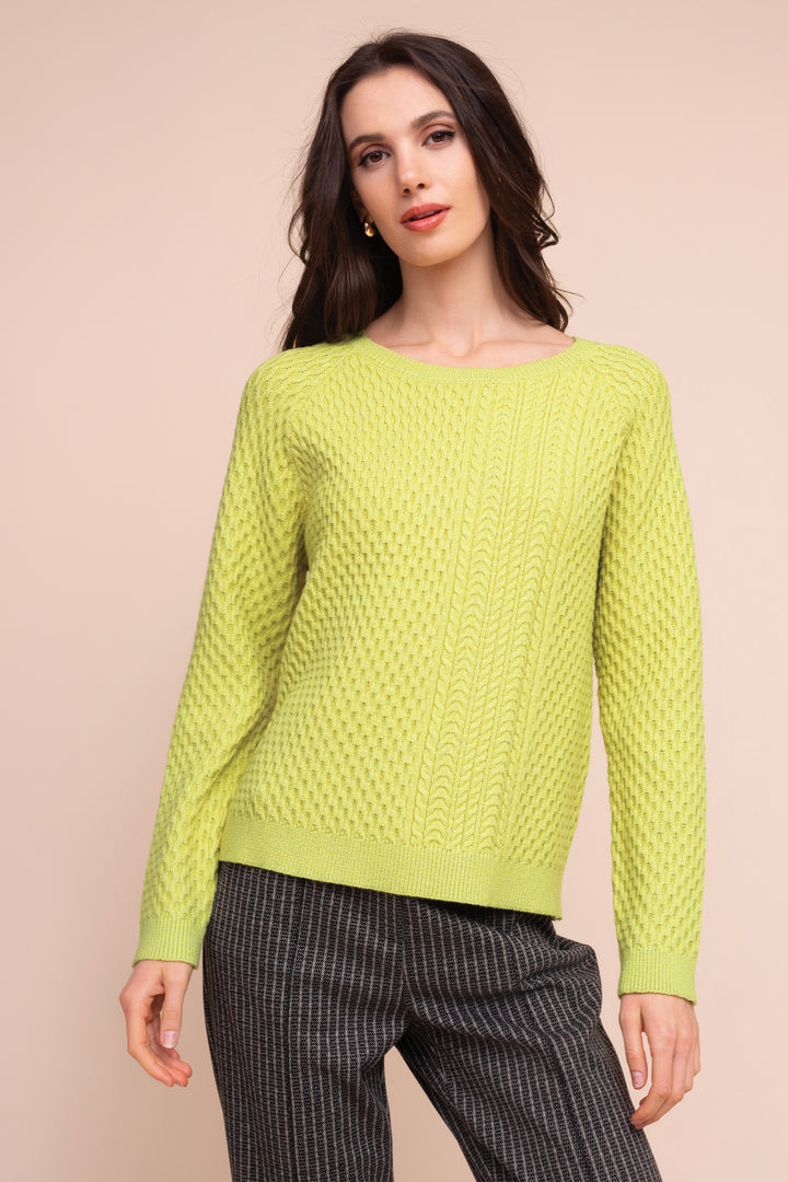A woman modeling a bright lime green knit sweater with a textured cable pattern, paired with dark gray pinstriped trousers, against a solid pale peach background.