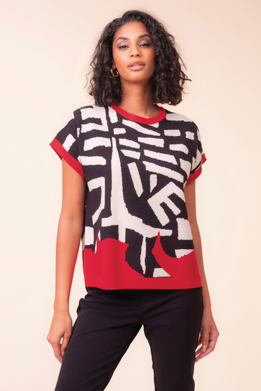 A woman modeling a short-sleeve knit top with an abstract black, white, and red geometric pattern, paired with black pants, against a solid beige background.