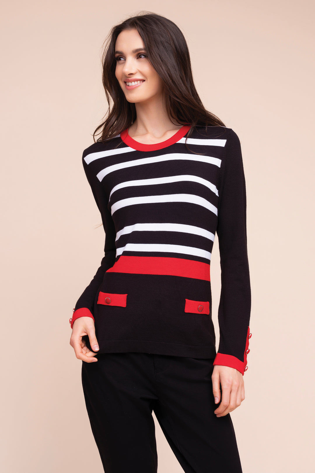A woman modeling a black and white horizontally striped long-sleeve sweater with red trim at the crew neck, cuffs, and hem, paired with black pants against a solid beige background.