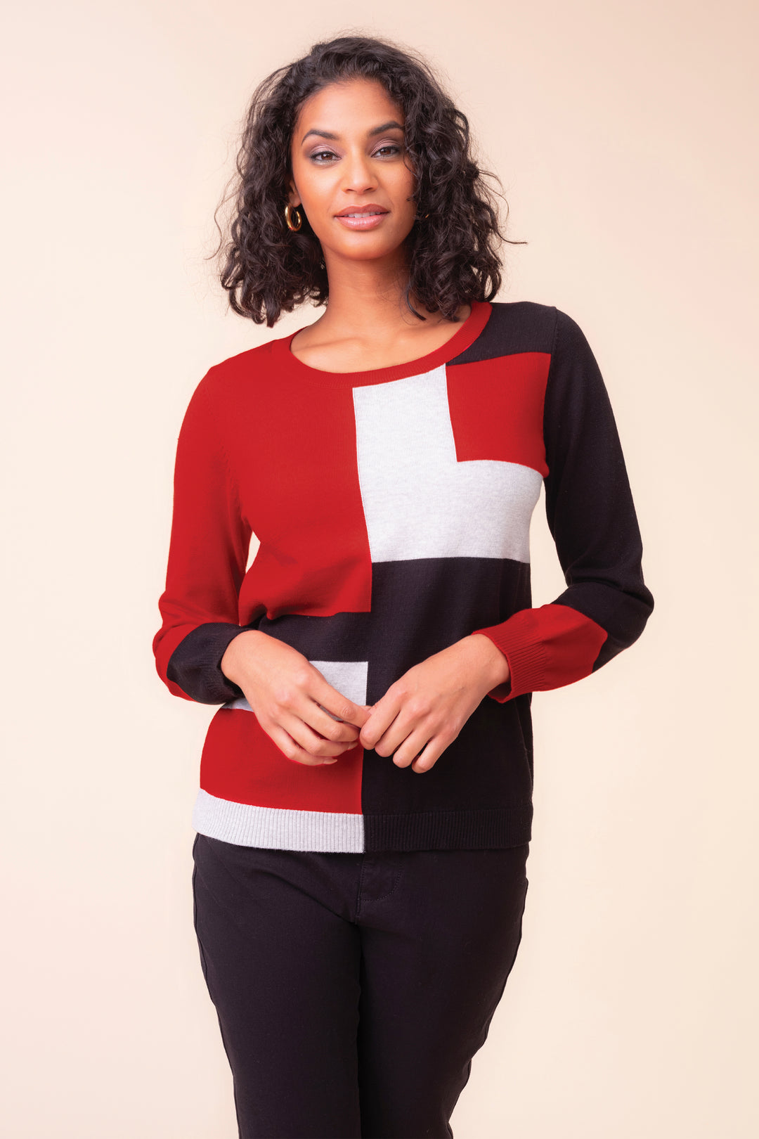 A woman modeling a color-block knit sweater with red, black, and white geometric panels, paired with black pants, against a solid beige background.