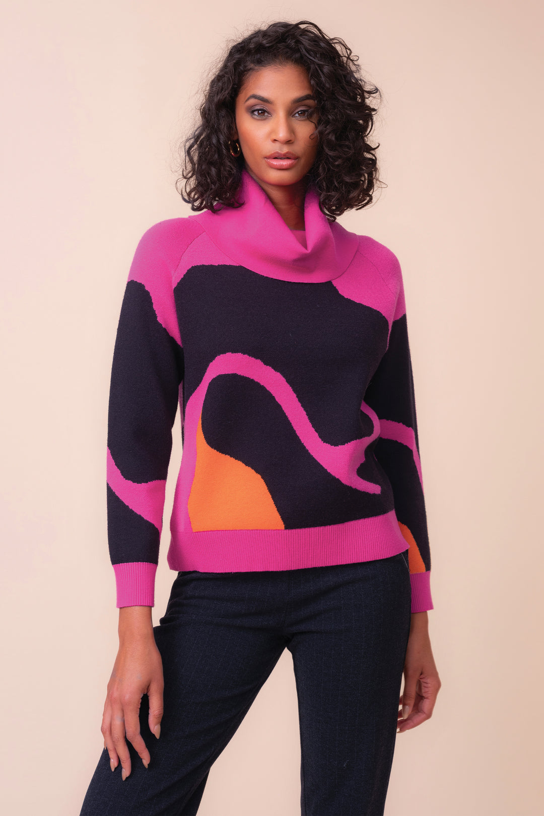 A woman modeling a long-sleeve cowl-neck sweater with a bold color-blocked pattern in pink, black, and orange, paired with dark jeans against a solid beige studio background.