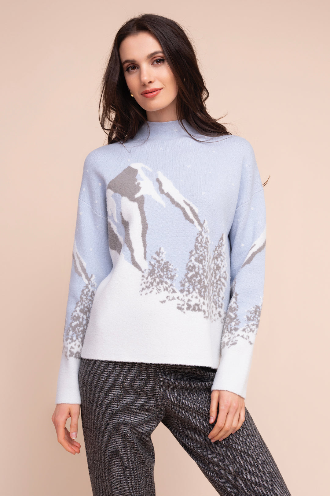 A woman modeling a light blue and white knit sweater with a mountain and tree pattern, paired with dark gray pants, against a solid beige background.