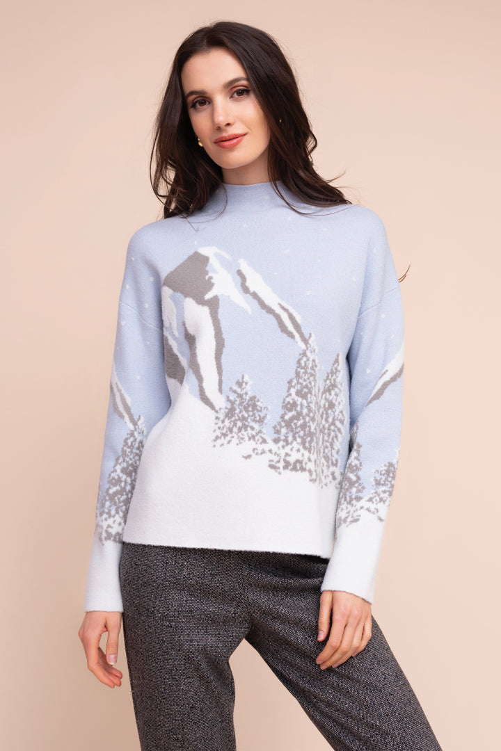 A woman modeling a light blue and white knit sweater with a mountain and tree pattern, paired with dark gray pants, against a solid beige background.