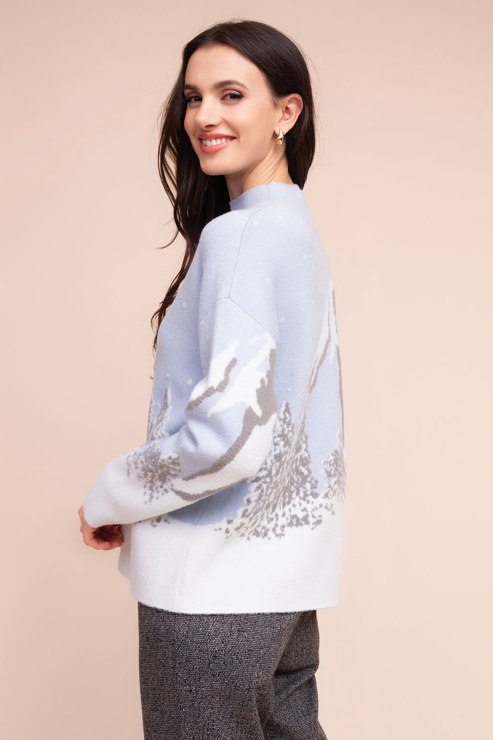 A woman modeling a light blue and white knit sweater with a gray mountain landscape pattern, paired with gray textured trousers, set against a solid beige background.