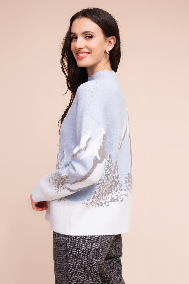 A woman modeling a light blue and white knit sweater with a gray mountain landscape pattern, paired with gray textured trousers, set against a solid beige background.
