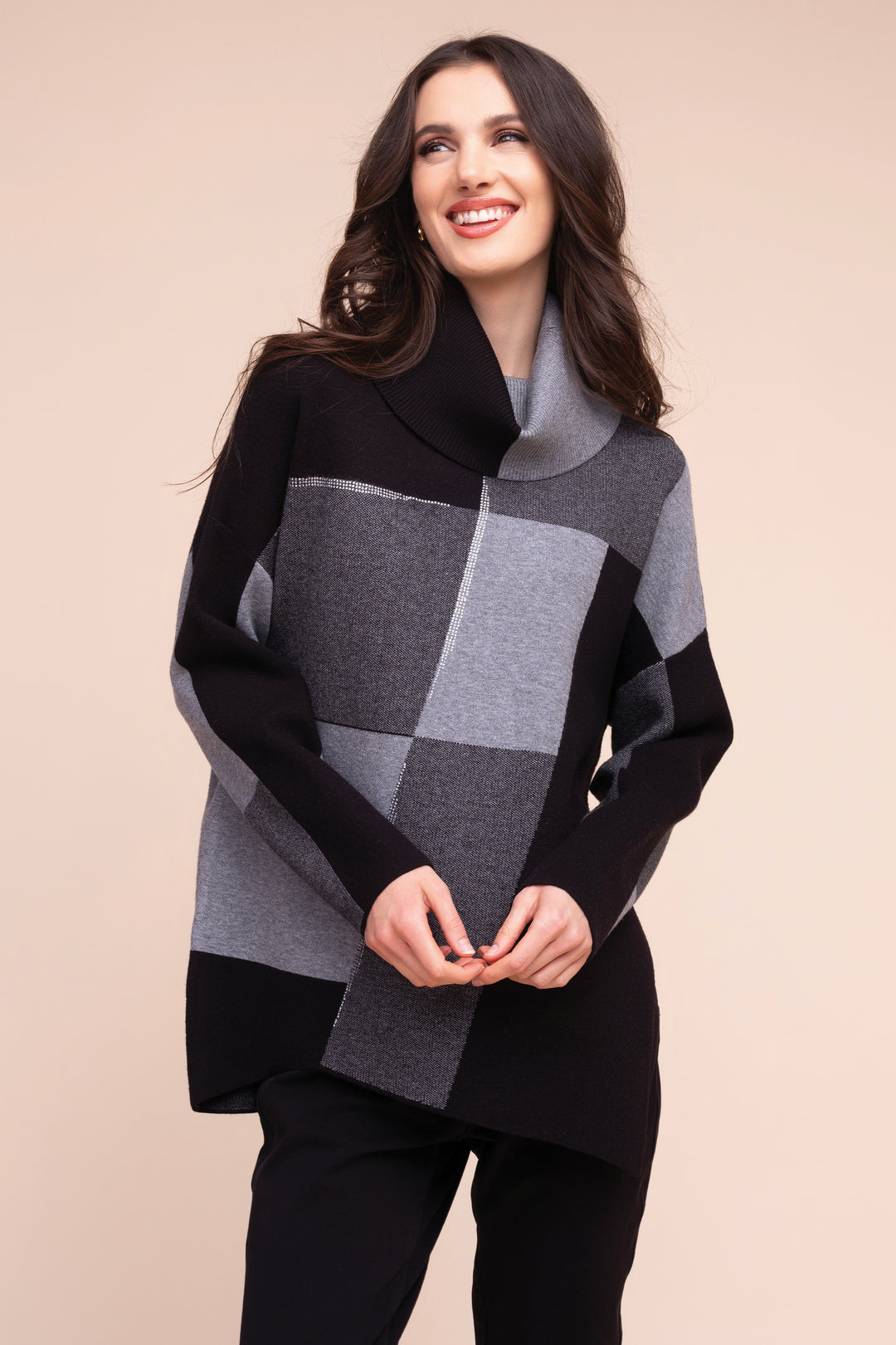 A woman modeling a black and gray patchwork cowl-neck sweater against a solid beige background.