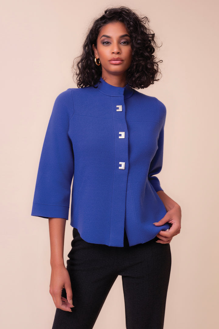 A woman modeling a royal blue cropped jacket with a high mock neck, three-quarter sleeves, and three decorative white geometric buttons down the front, paired with black pants against a solid beige background.