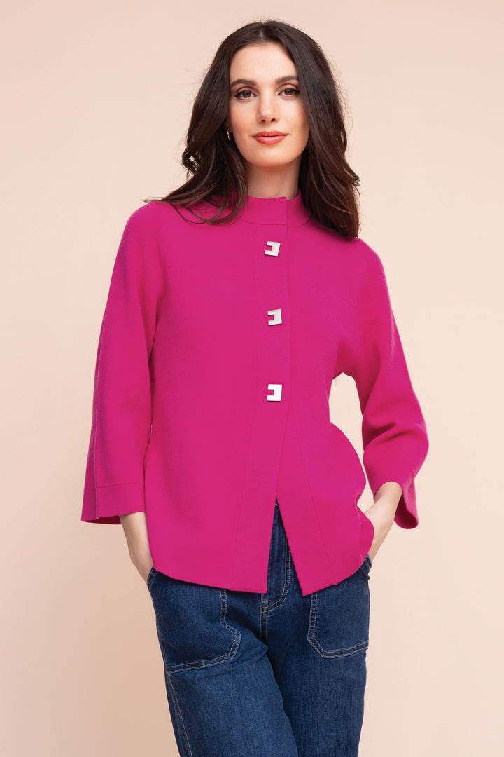 A woman modeling a bright magenta pink long-sleeve top with three decorative white square buttons down the front, paired with blue jeans, against a solid light beige background.