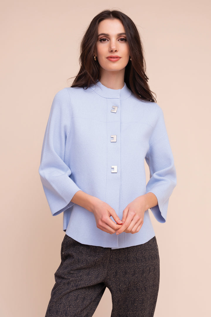 A woman modeling a light blue high-neck blouse with three decorative square buttons down the front and flared three-quarter sleeves, paired with dark gray textured trousers, against a solid beige background.