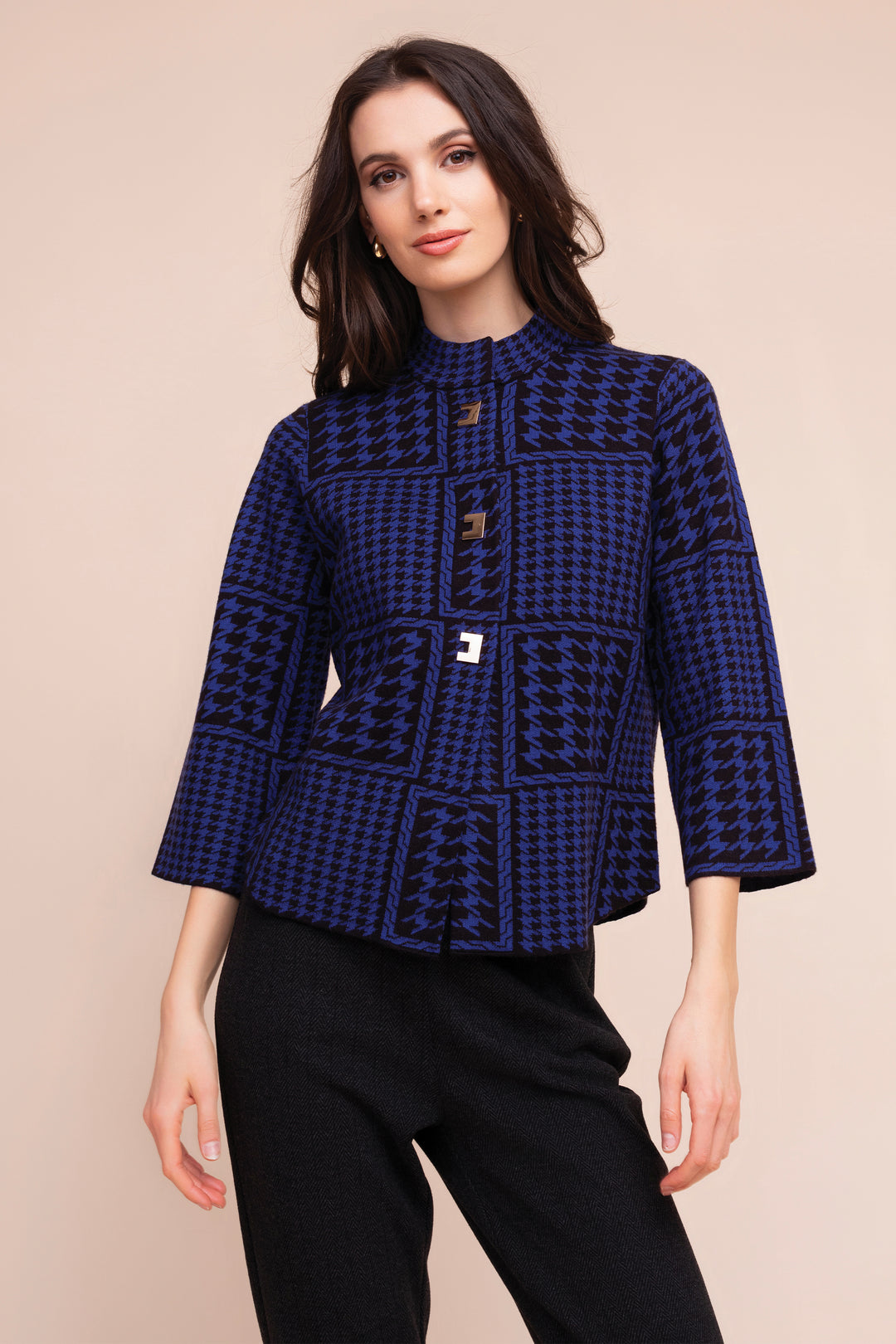 A woman modeling a navy blue and black patterned jacket with a mock neck and three-quarter sleeves, paired with black pants, against a solid beige background.