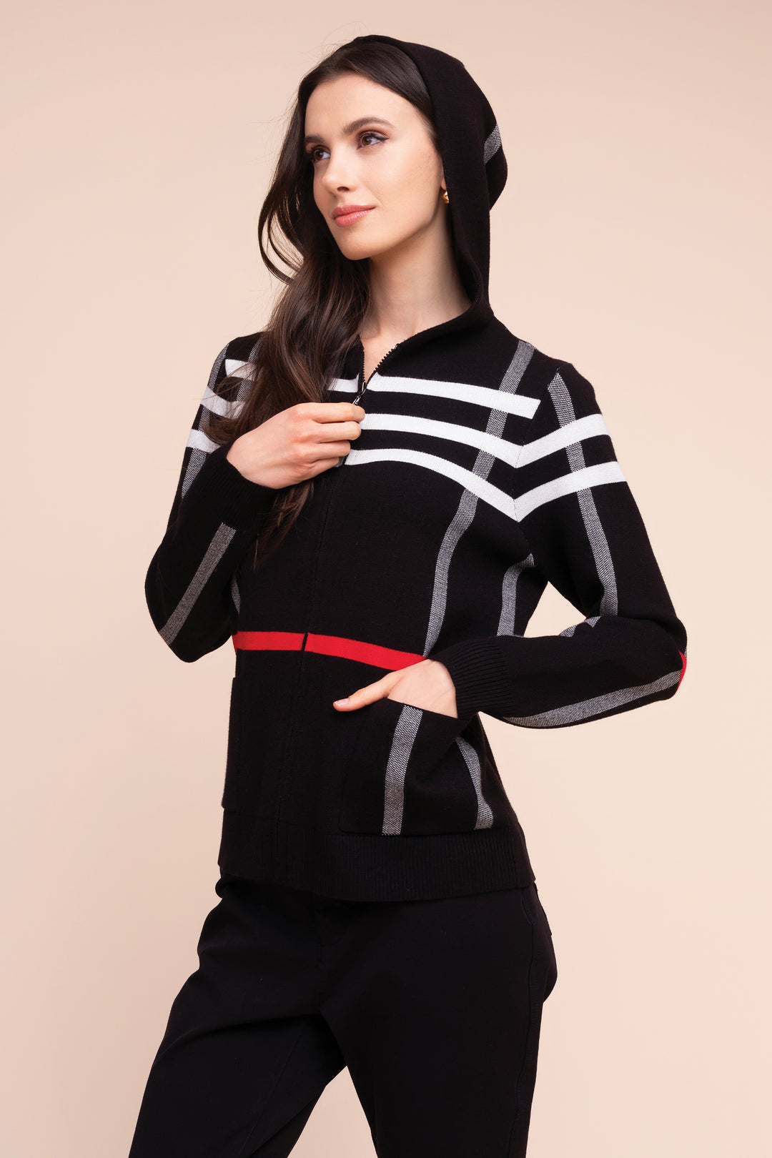 A woman modeling a black zip-up hoodie with white geometric stripe patterns, gray vertical accents, and a red waist detail, paired with black pants, against a solid beige background.