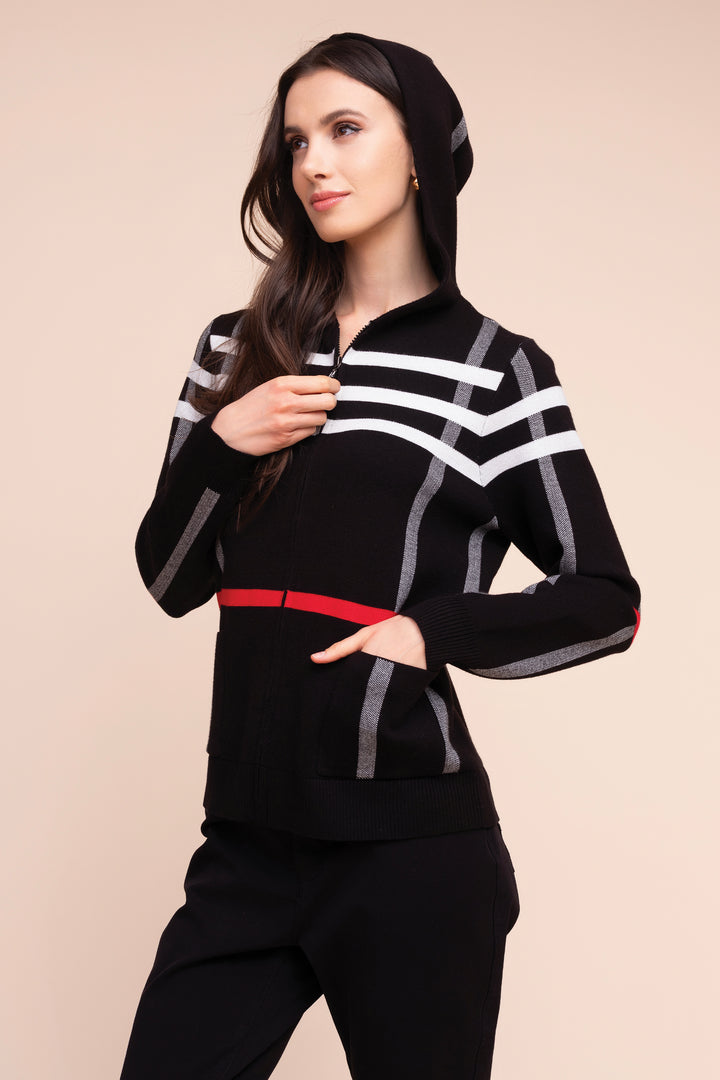 A woman modeling a black zip-up hoodie with white geometric stripe patterns, gray vertical accents, and a red waist detail, paired with black pants, against a solid beige background.