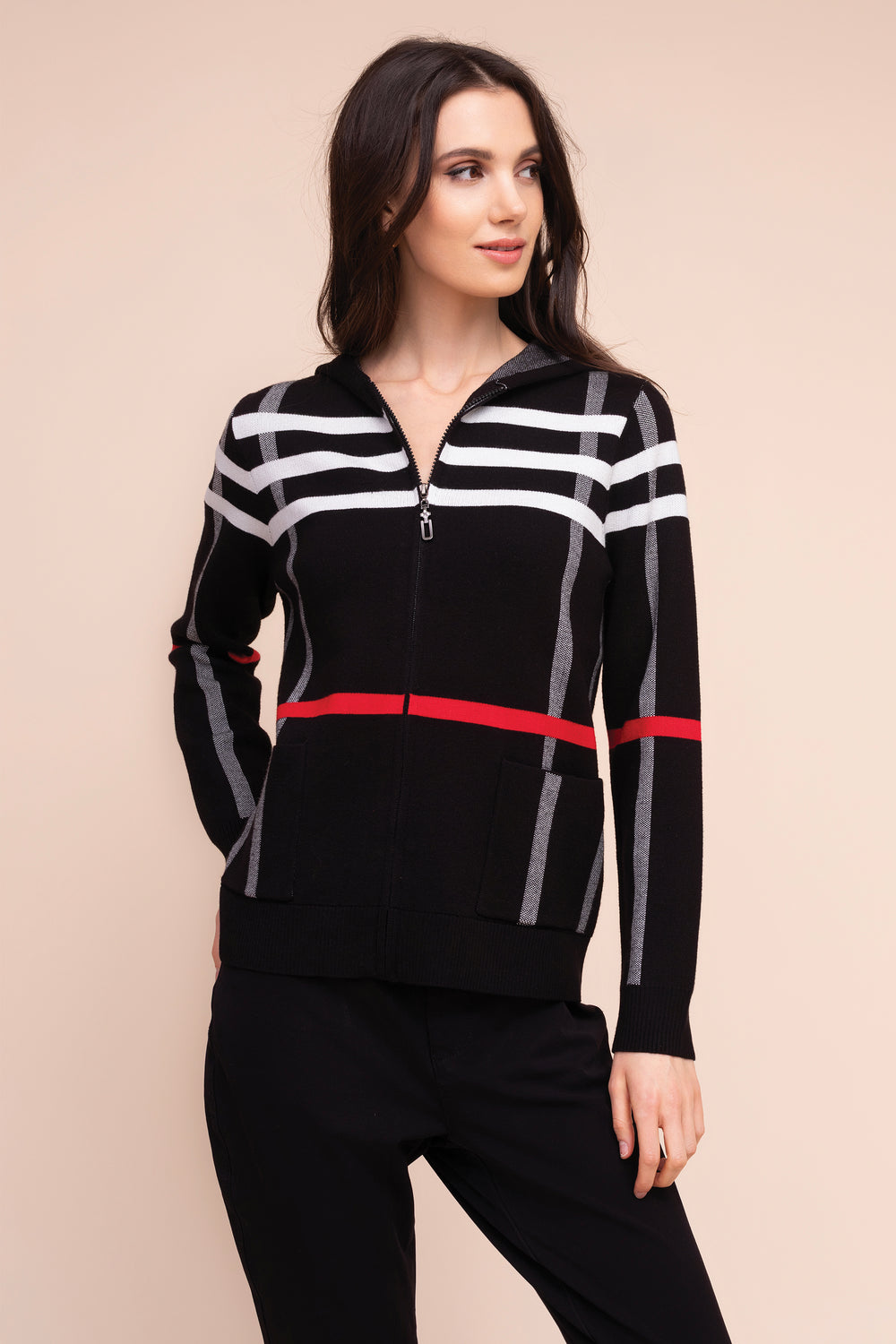 A woman modeling a black zip-up knit sweater with white and gray horizontal stripes on the chest and sleeves, accented by a red horizontal stripe at the waist, paired with black pants against a solid beige background.