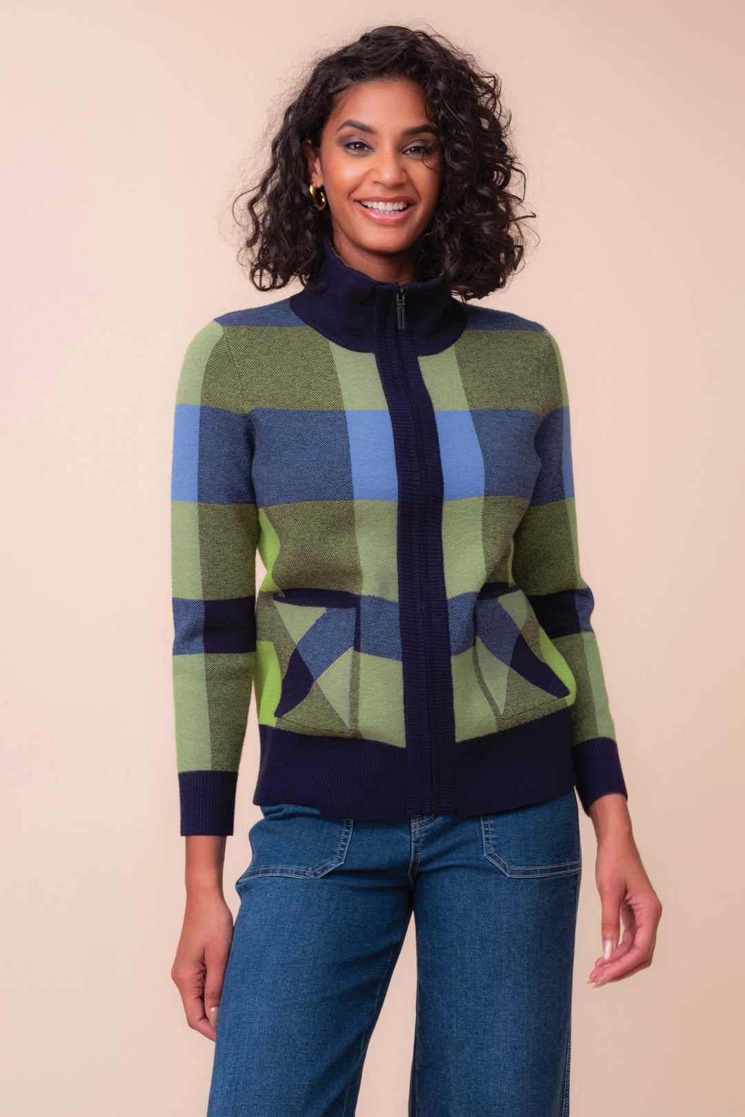 A woman modeling a colorful knit zip-up cardigan sweater with a geometric plaid pattern in shades of green, blue, and navy, paired with blue jeans against a solid beige background.