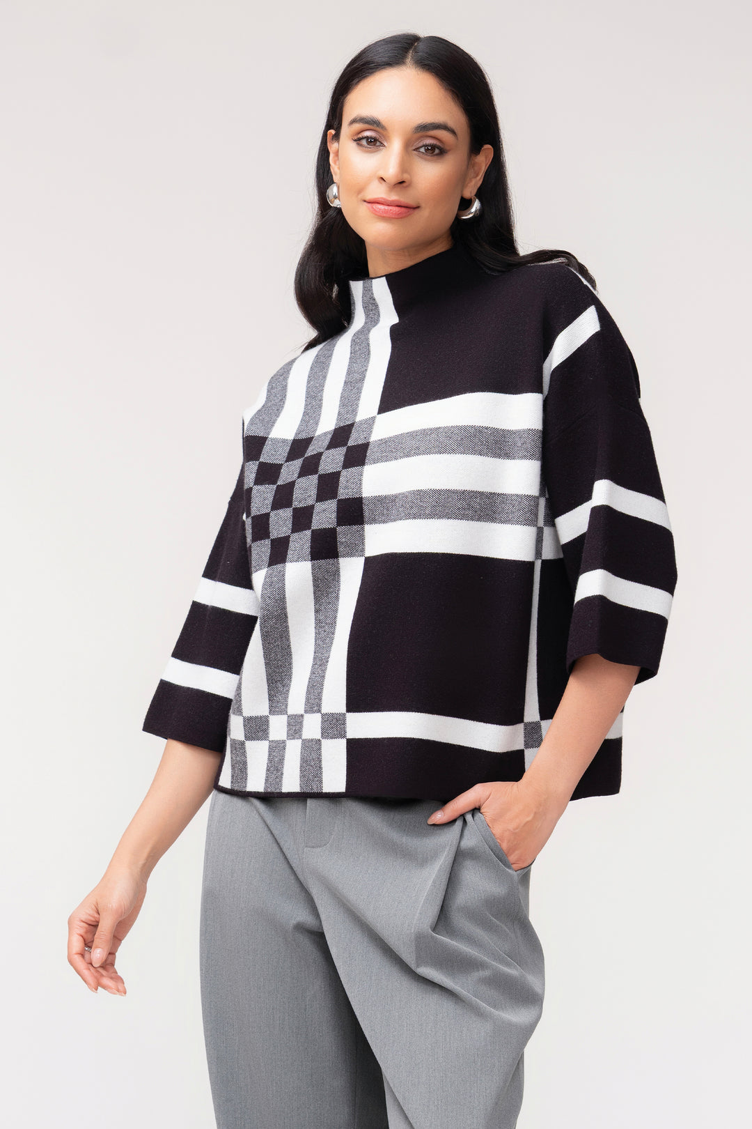 A woman modeling a black, white, and gray striped knit mock-neck top with a checkerboard pattern on the front, paired with gray trousers, against a light gray studio background.