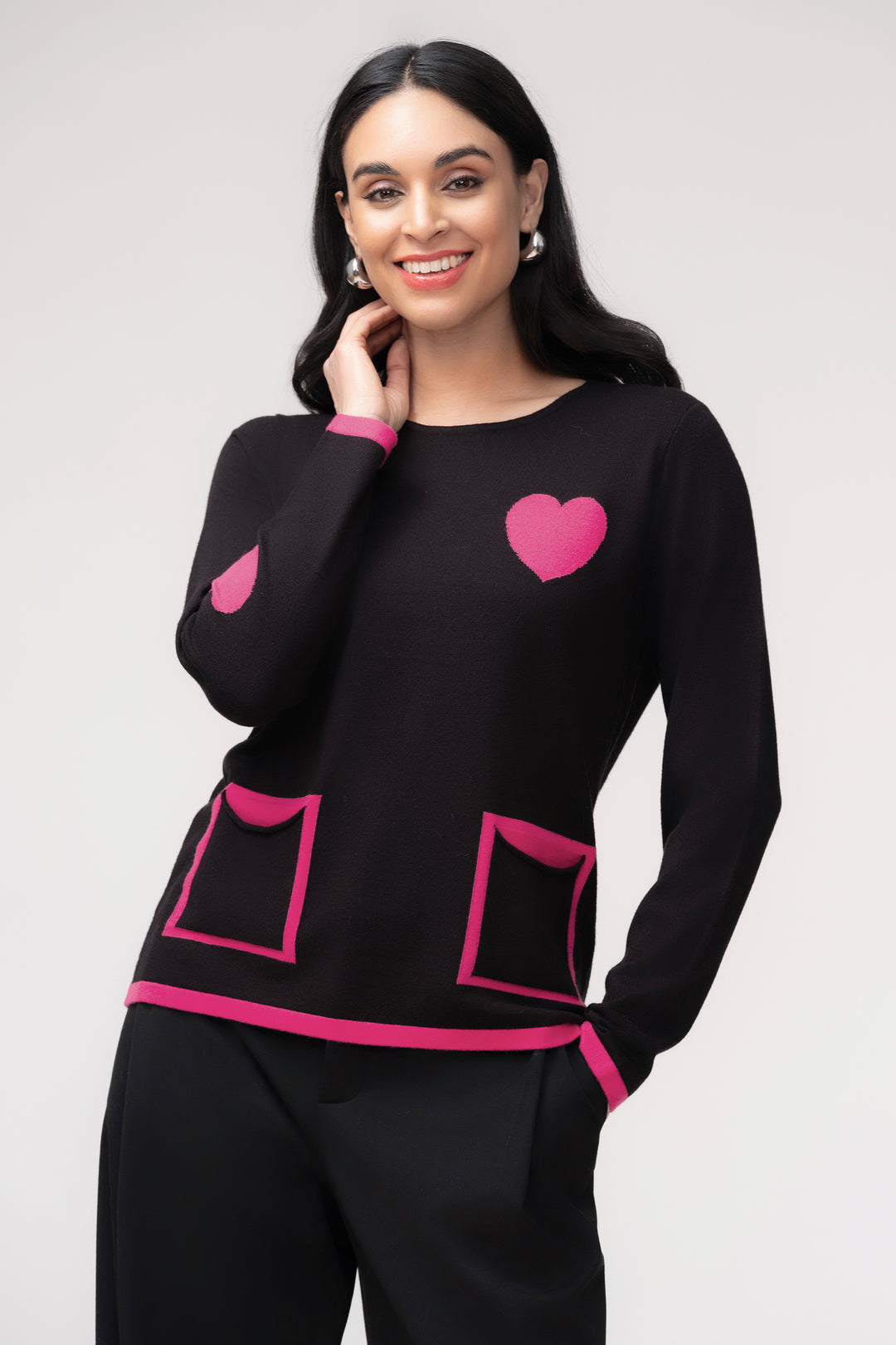 A woman modeling a black knit sweater with bright pink heart and pocket details, paired with black pants, against a light gray studio background.